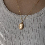 A gold oval locket necklace with a chain, displayed on a person wearing a white knit top.