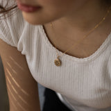 A gold oval locket necklace with a chain, displayed on a person wearing a white knit top.